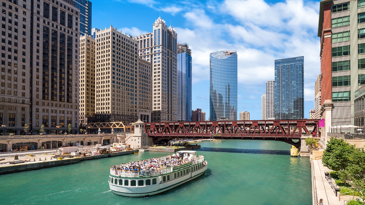 Chicago River cruise boat passing under a bridge with city skyscrapers in the background.