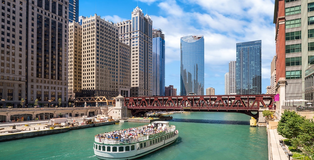 Chicago River cruise boat passing under a bridge with city skyscrapers in the background.