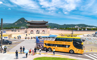 Yellow city tour bus near Gwanghwamun Gate, Seoul, with tourists and mountain backdrop.