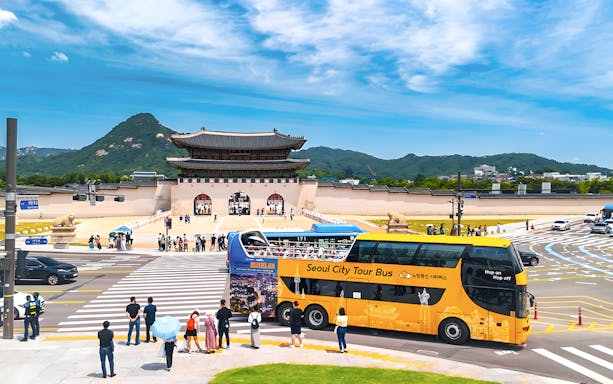 Yellow city tour bus near Gwanghwamun Gate, Seoul, with tourists and mountain backdrop.