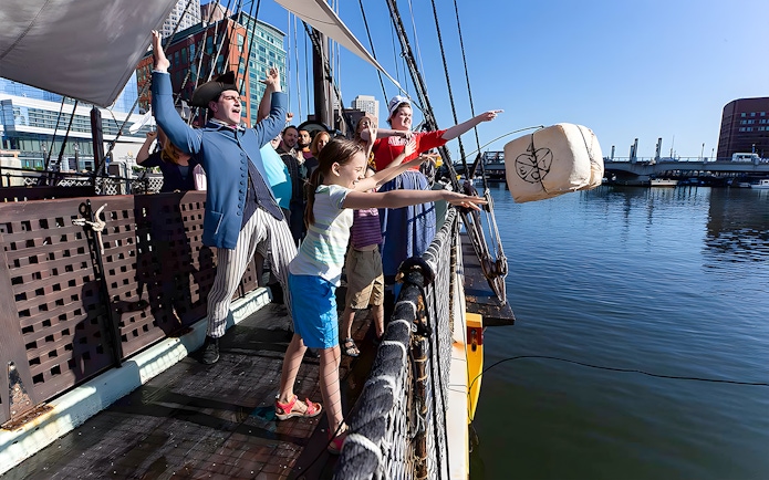 Tourists reenact Boston Tea Party by throwing tea chest from ship into harbor.
