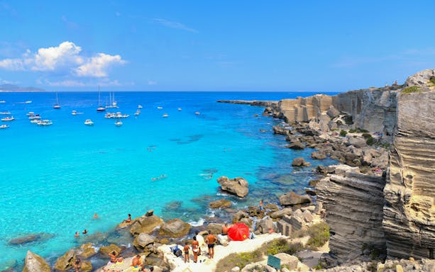 Cala Rossa beach in Sicily with turquoise waters, boats, and rocky cliffs.