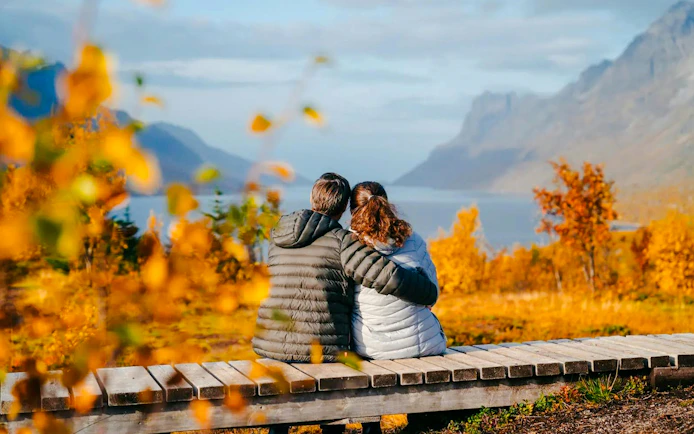 Couple sitting on a bench overlooking Sommarøy Fjord with autumn foliage, Tromsø.