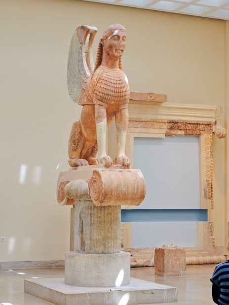 Sphinx of Naxos statue in Delphi museum, Greece, with visitors observing.
