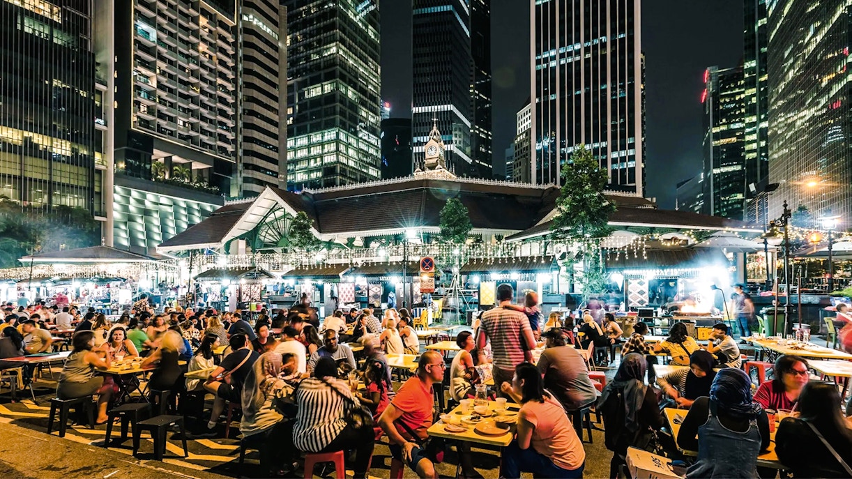 Crowded outdoor dining at La Pau Sat food market, Singapore, with skyscrapers in the background.