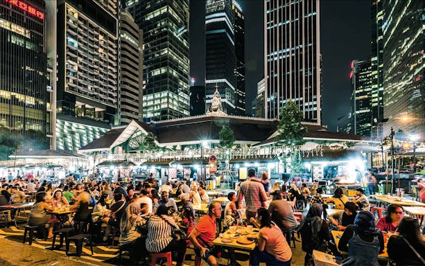 Crowded outdoor dining at La Pau Sat food market, Singapore, with skyscrapers in the background.