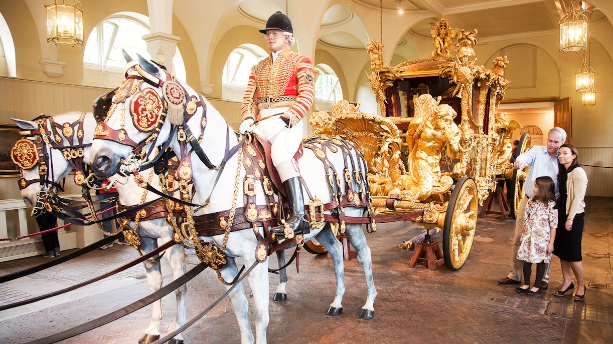 Royal Mews at Buckingham Palace with ornate carriage and horses, guide explaining to visitors.