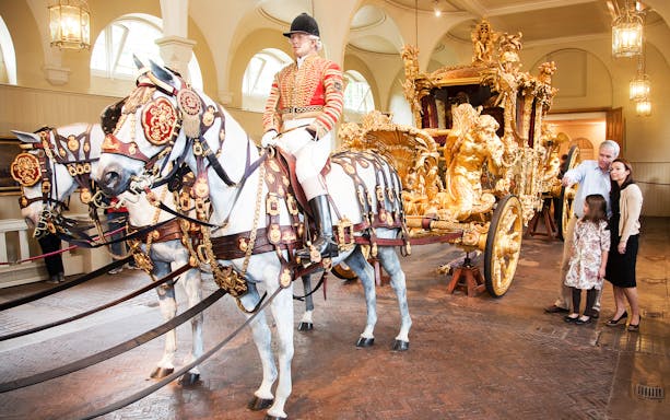 Royal Mews at Buckingham Palace with ornate carriage and horses, guide explaining to visitors.