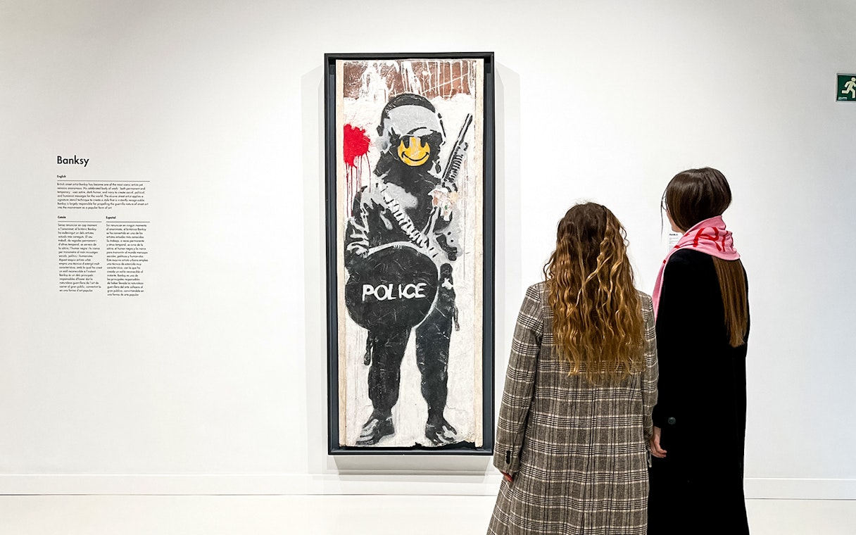 Tourists viewing Banksy artwork of a police figure with a smiley face.