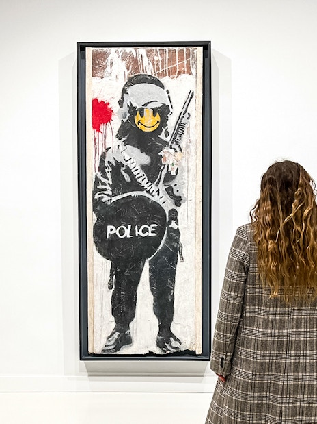 Tourists viewing Banksy artwork of a police figure with a smiley face.