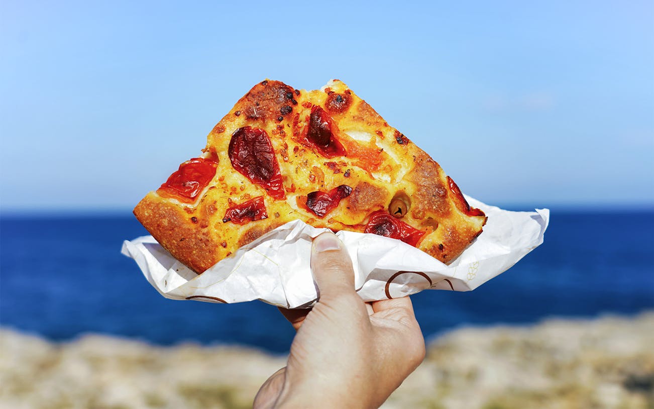 Focaccia slice with tomatoes held by hand, Polignano a Mare coastline in background.