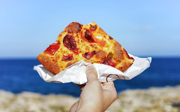 Focaccia slice with tomatoes held by hand, Polignano a Mare coastline in background.