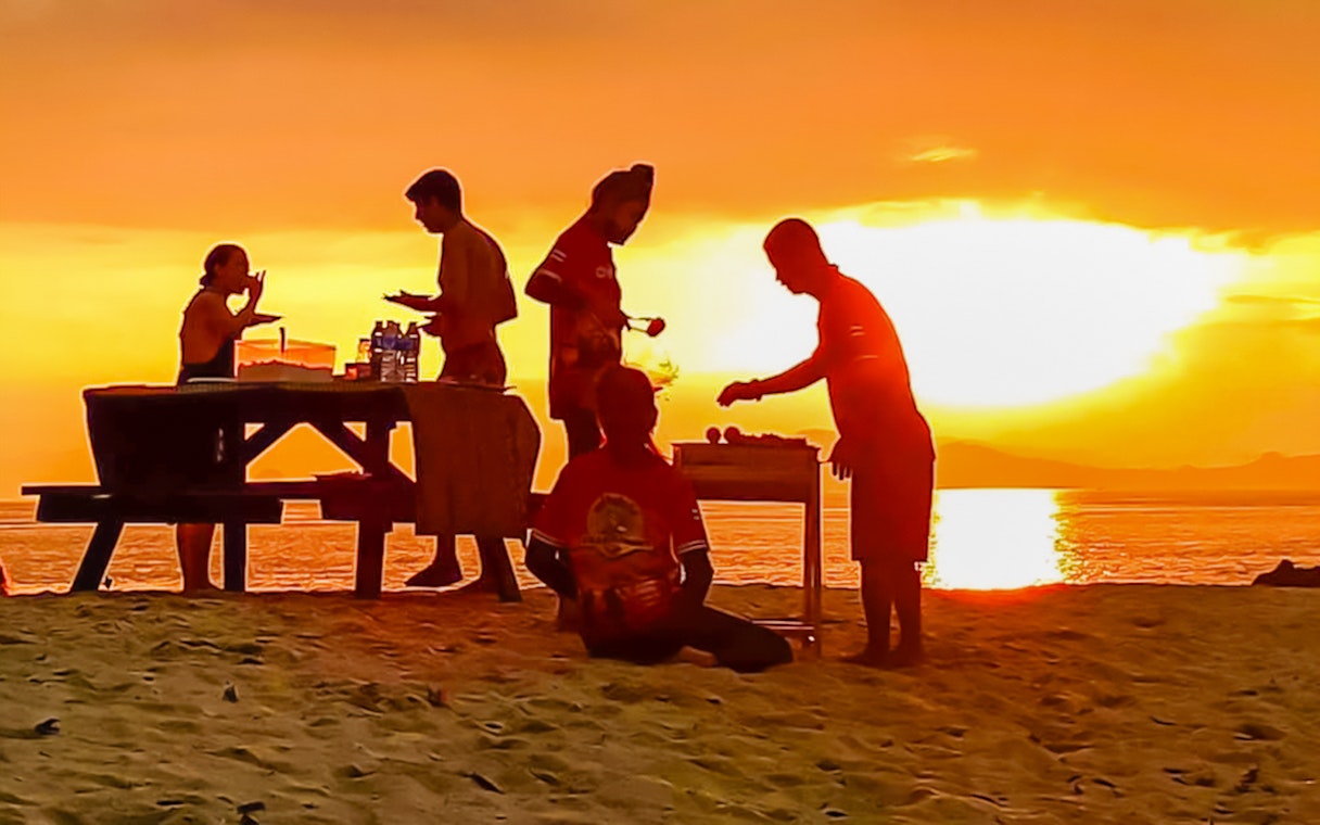 Group enjoying a beach barbeque at sunset with ocean view.