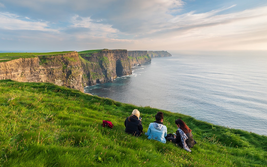 Visitors sitting near the edge of the Cliffs of Moher, Ireland, overlooking the ocean.