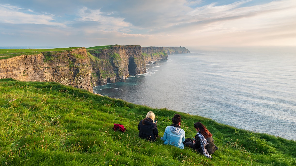 Visitors sitting near the edge of the Cliffs of Moher, Ireland, overlooking the ocean.