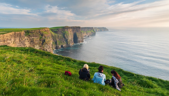 Visitors sitting near the edge of the Cliffs of Moher, Ireland, overlooking the ocean.