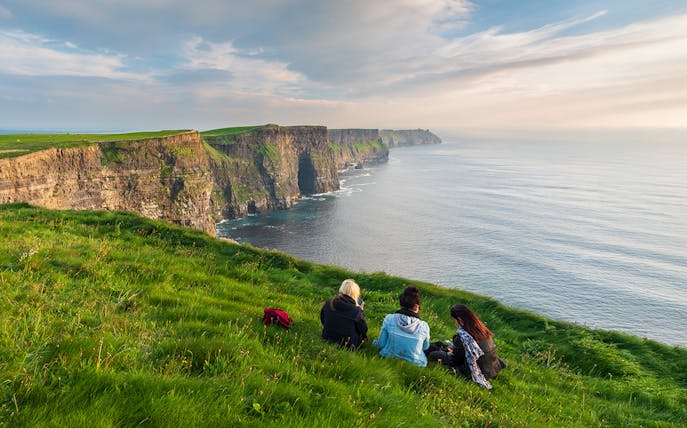 Visitors sitting near the edge of the Cliffs of Moher, Ireland, overlooking the ocean.