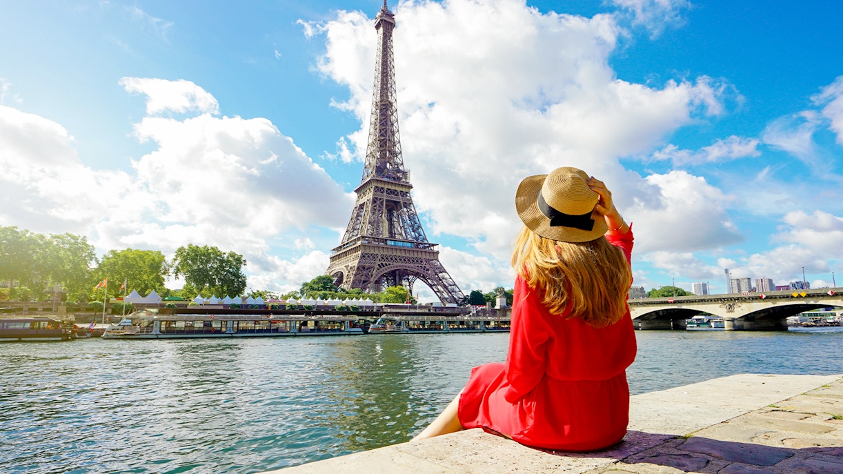 Person in red dress sitting by the Seine River with Eiffel Tower in background, Paris.