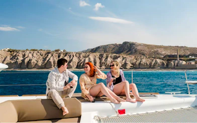 Guests relaxing on a catamaran cruise with drinks, scenic coastal cliffs in the background.