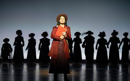 Performer in period costume on stage with silhouetted figures in the background, related to Suffs.