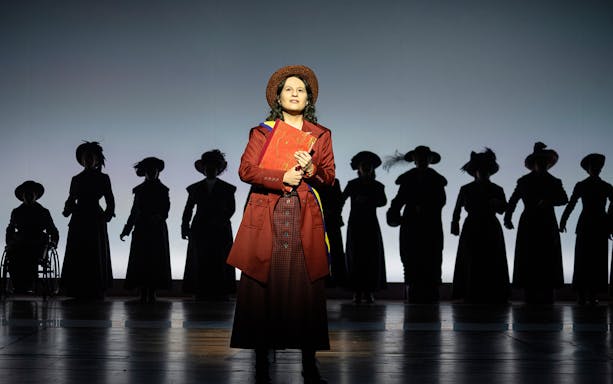 Performer in period costume on stage with silhouetted figures in the background, related to Suffs.