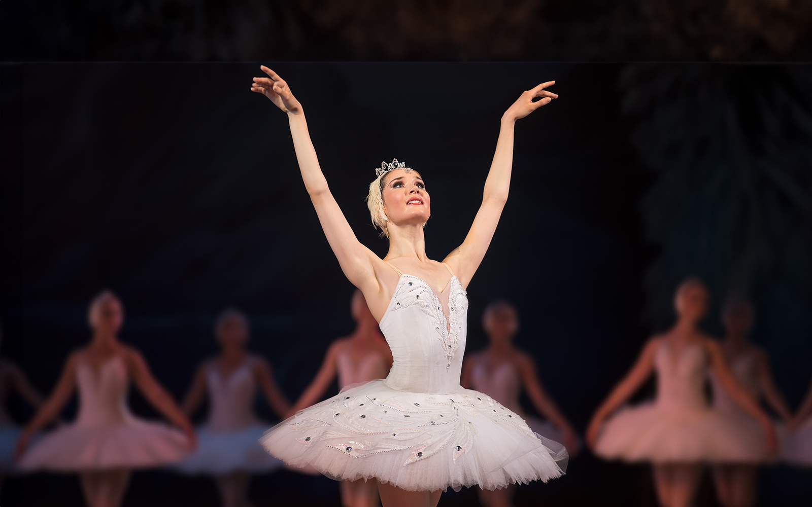 Ballet dancer performing on stage in a white tutu with arms raised.