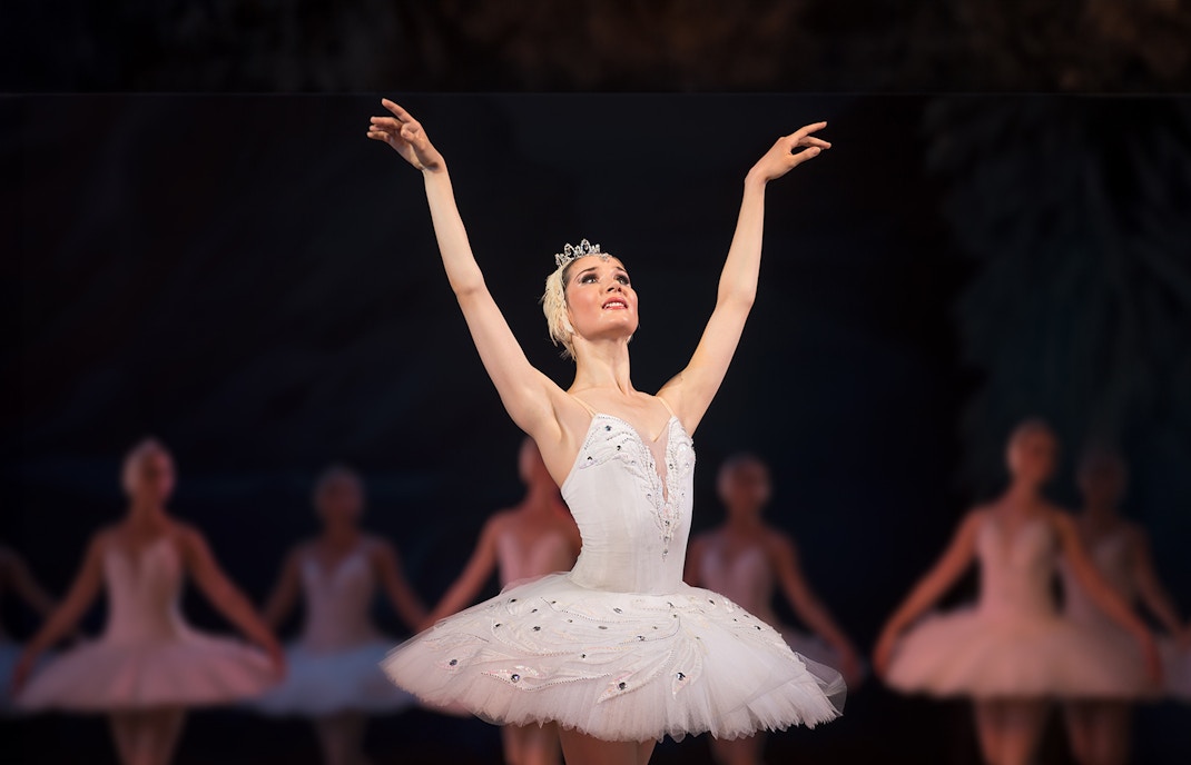Ballet dancer performing on stage in a white tutu with arms raised.