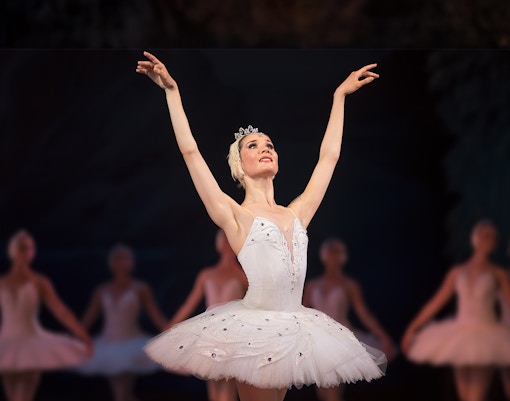 Ballet dancer performing on stage in a white tutu with arms raised.