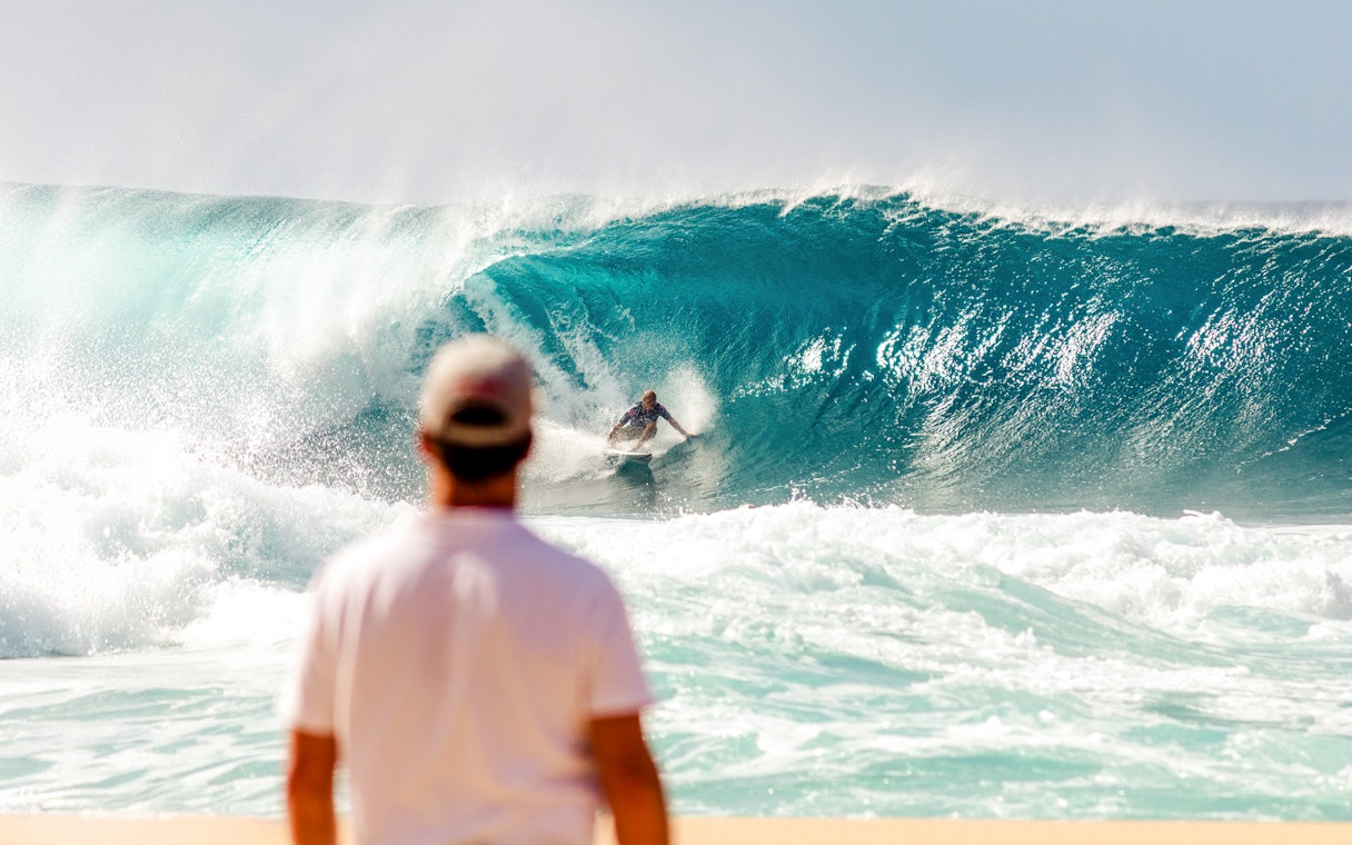 Tourist watching a surfer ride a large wave at the beach.
