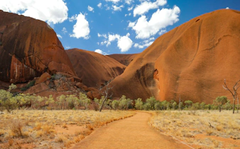 Uluru rock formation with walking path and trees under a blue sky, Alice Springs tour.
