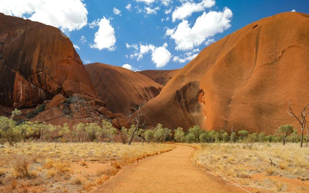 Uluru rock formation with walking path and trees under a blue sky, Alice Springs tour.