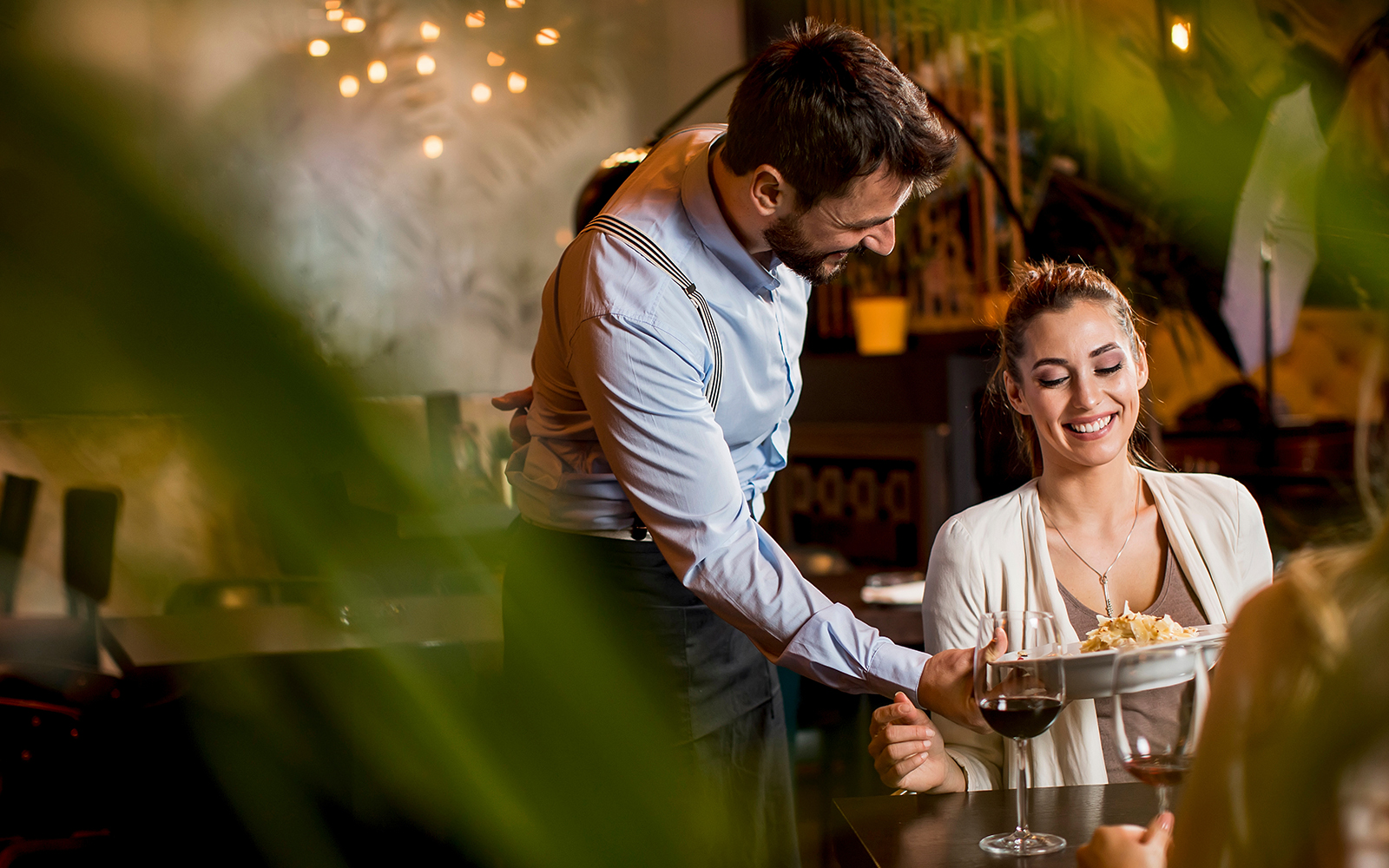 Waiter serving a dish to a smiling woman at a restaurant table.