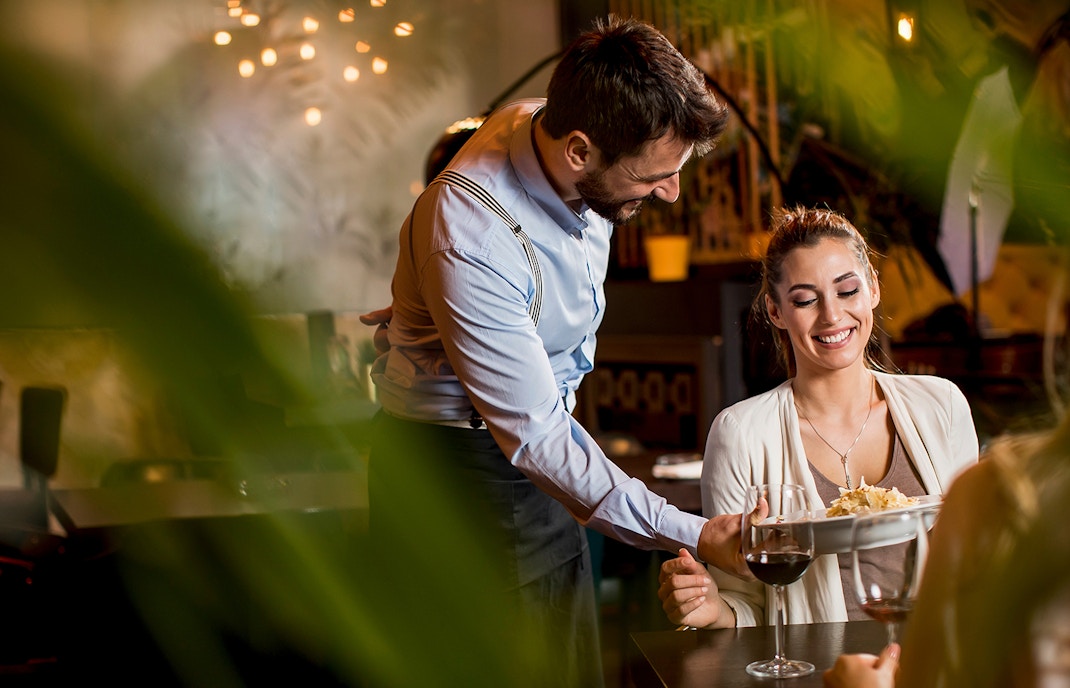 Waiter serving a dish to a smiling woman at a restaurant table.