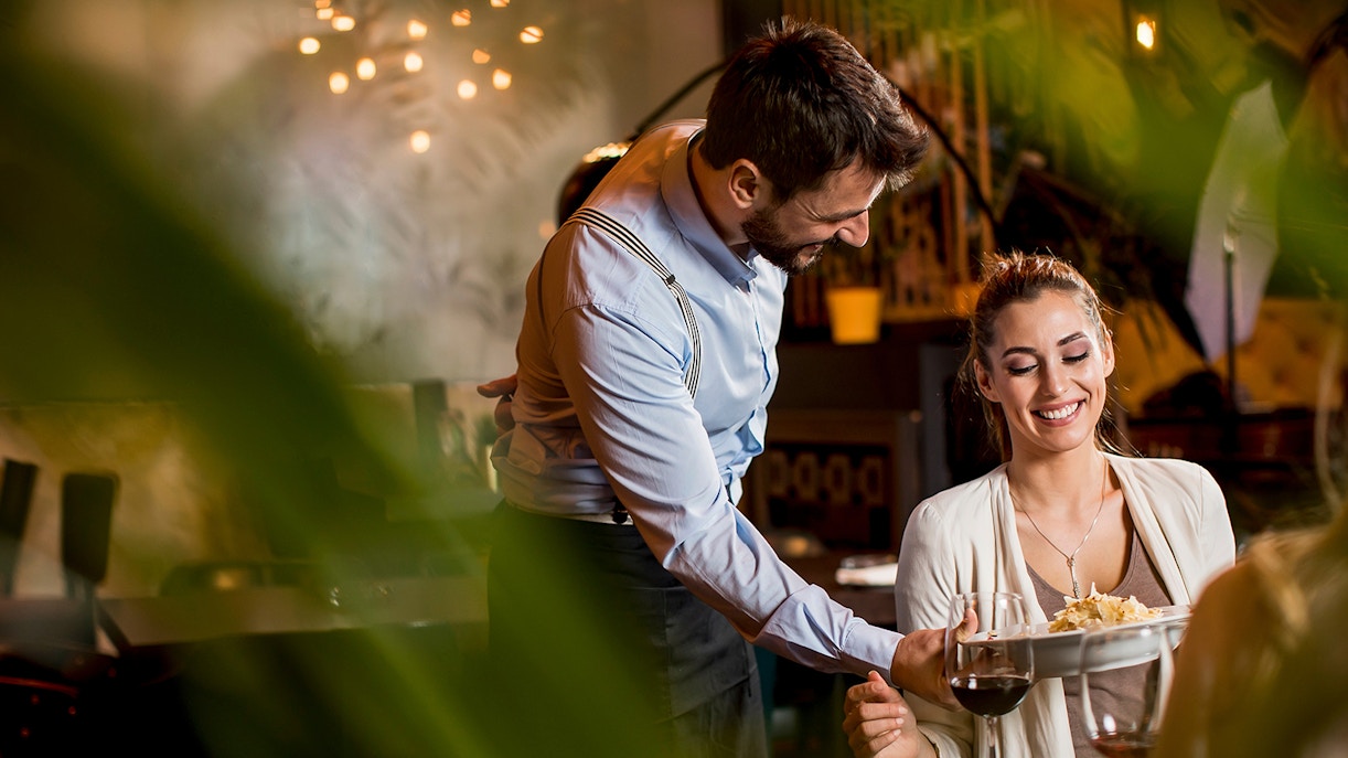 Waiter serving a dish to a smiling woman at a restaurant table.
