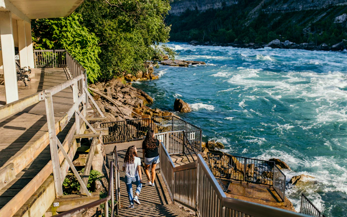 Visitors descending stairs towards white water platform by a river.