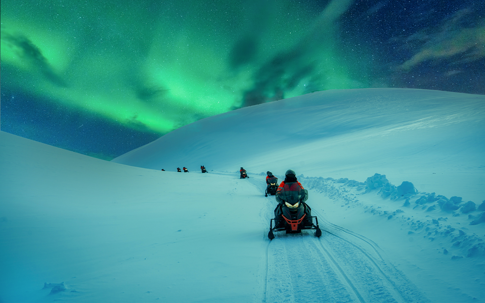 Snowmobiles on snowy mountain trail under aurora borealis.