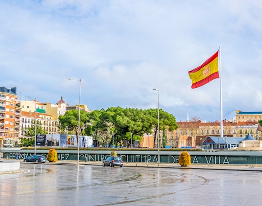 Spain flag at Plaza de Colón, Madrid, with surrounding buildings and trees.