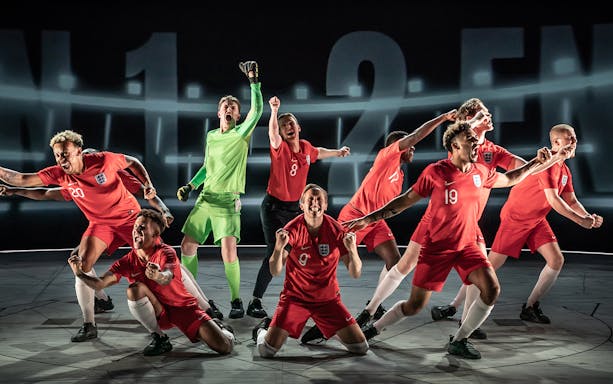 Football players in red England jerseys celebrating on stage, "Dear England" play scene.