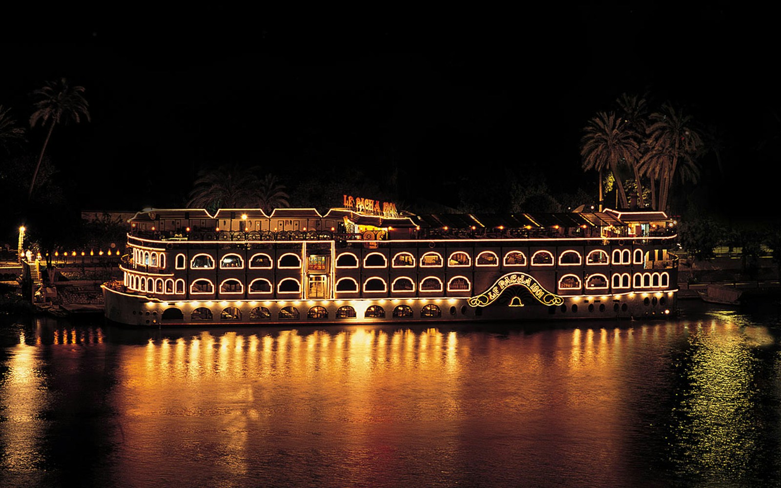 Cruise ship illuminated at night on the Nile River, Egypt, with palm trees in the background.