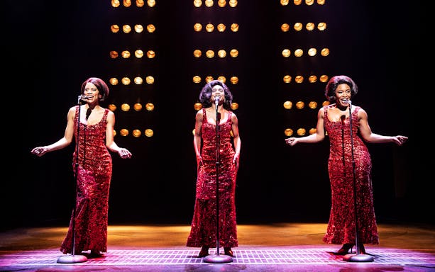 Performers in red dresses singing on stage during "Ain't Too Proud" musical.