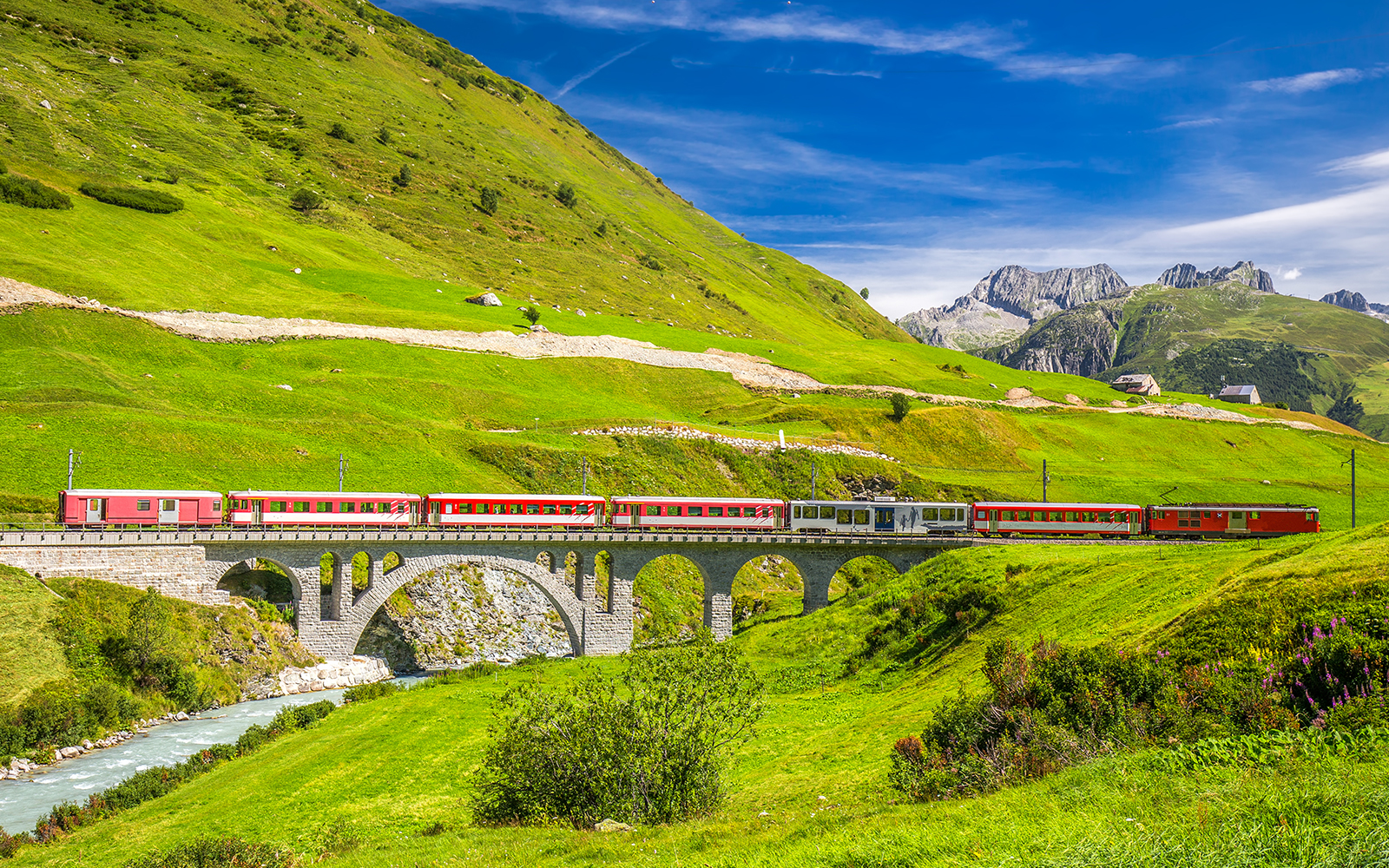 Aerial view of red passenger train traveling across a bridge over a waterfall, surrounded by colorful autumn forest in the swiss alps.