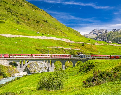 Aerial view of red passenger train traveling across a bridge over a waterfall, surrounded by colorful autumn forest in the swiss alps.