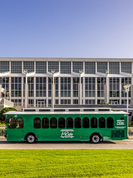 Green hop-on hop-off trolley in front of a large convention center with palm trees.