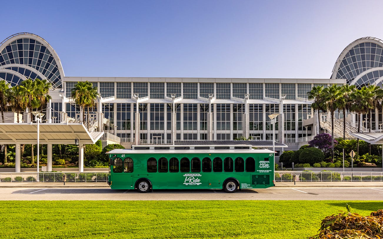 Green hop-on hop-off trolley in front of a large convention center with palm trees.