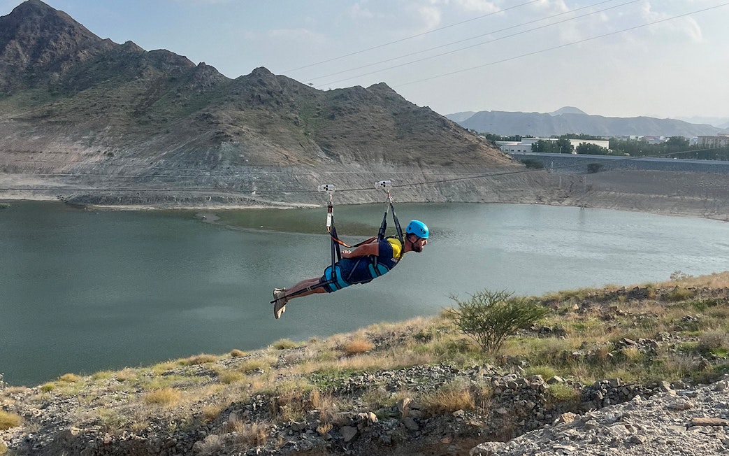 Person ziplining over a lake at Fujairah Adventure Park with mountains in the background.