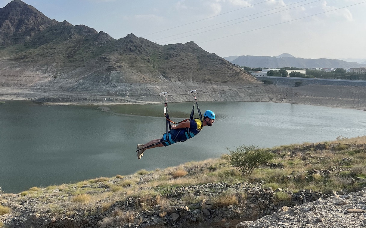 Person ziplining over a lake at Fujairah Adventure Park with mountains in the background.