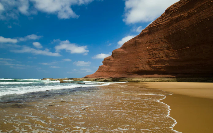 Huge red cliffs on Legzira beach, Morocco, with waves hitting the shore.