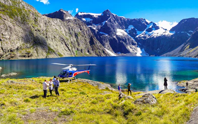 Helicopter landing by a lake in Milford Sound with people exploring the scenic mountain landscape.