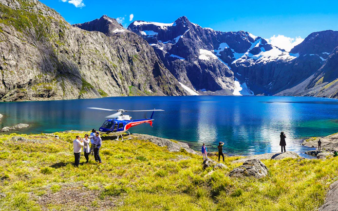 Helicopter landing by a lake in Milford Sound with people exploring the scenic mountain landscape.