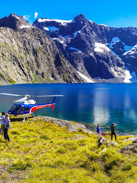 Helicopter landing by a lake in Milford Sound with people exploring the scenic mountain landscape.